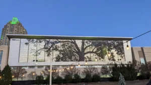 Raleigh Convention Center Building exterior featuring a large tree mural, illuminated at dusk, with surrounding greenery and string lights, symbolizing local community engagement and aesthetics in Raleigh.