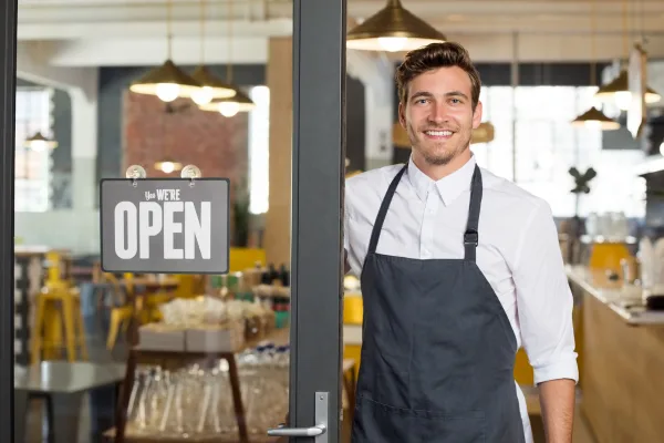 Smiling man in an apron standing at a café entrance holding an "Open" sign, representing small local shops ready to serve customers.